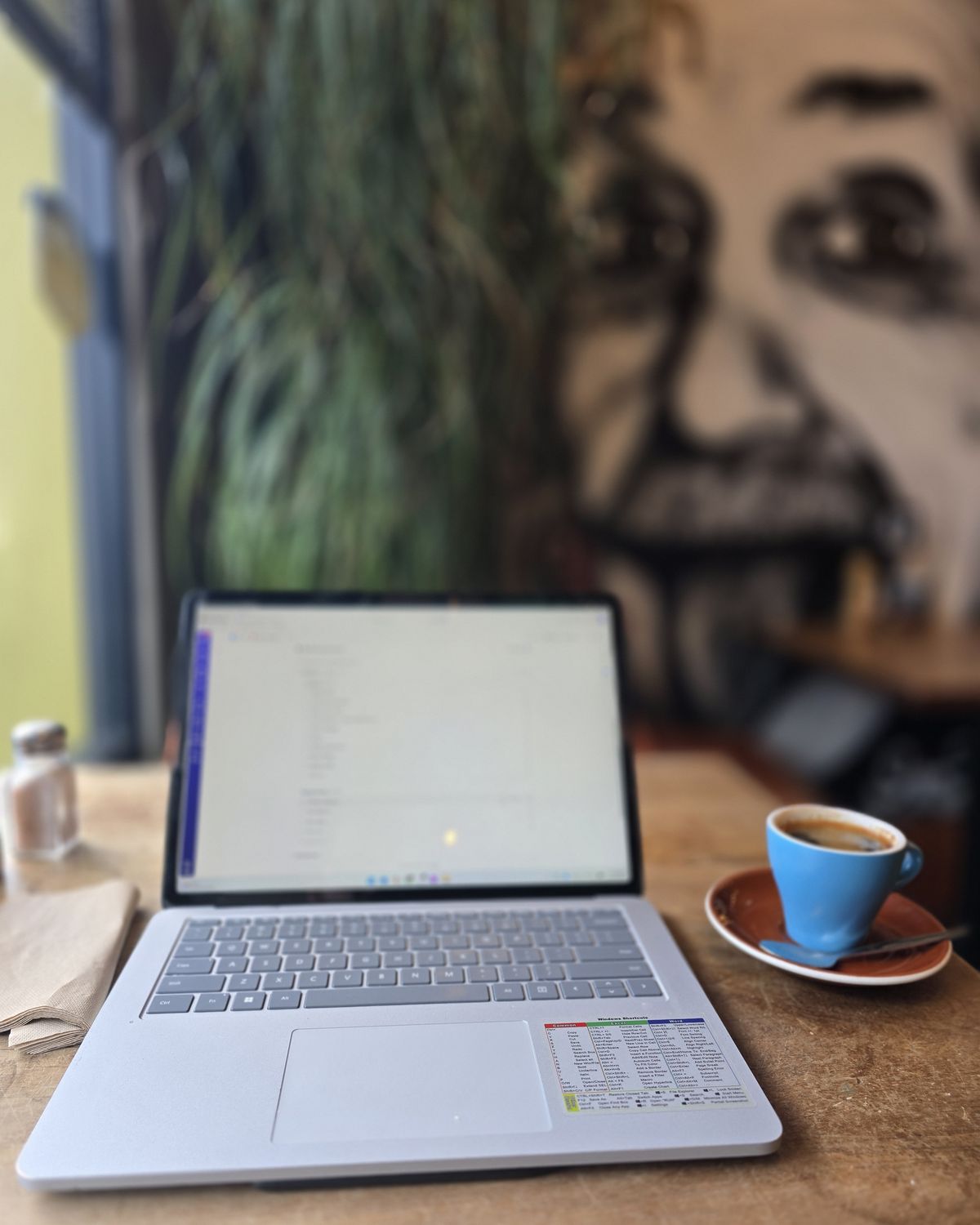 A laptop open on a wooden café table beside a small blue coffee, with a large mural of Albert Einstein on the wall behind.