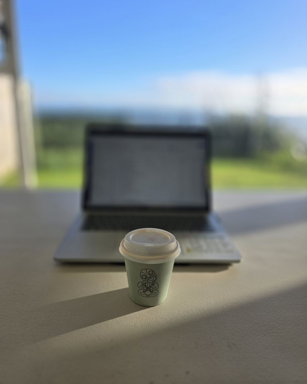 A takeaway coffee in the morning sun on a desk, with a laptop and green hills blurred behind.